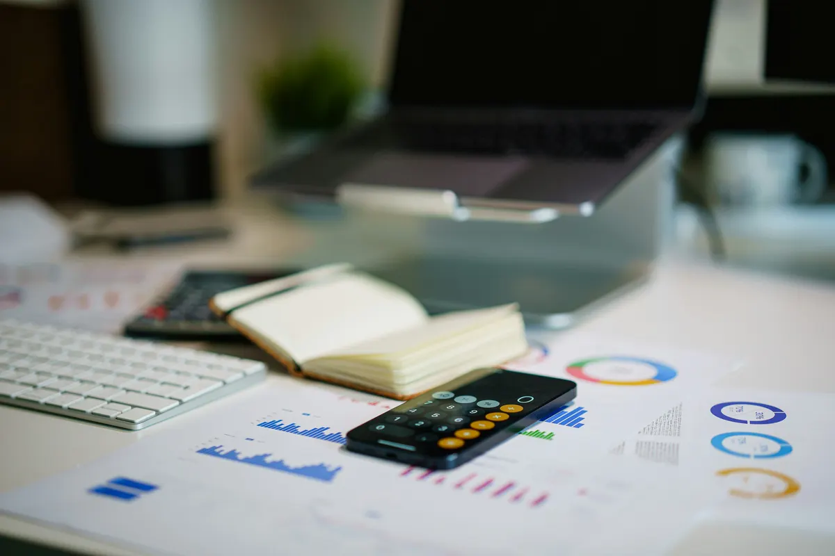 A calculator, notebook and phone on a desk beside printed charts